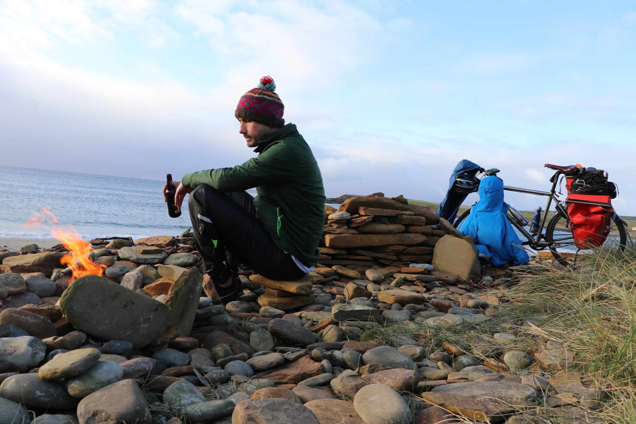 Person wearing a beanie hat sat on a pebble beach holding a bottle beside a fire, a bicycle stands in the background