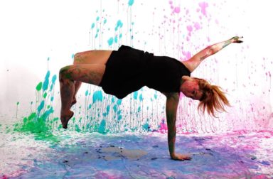 A woman supports her weight on one arm mid-flip covered in paint