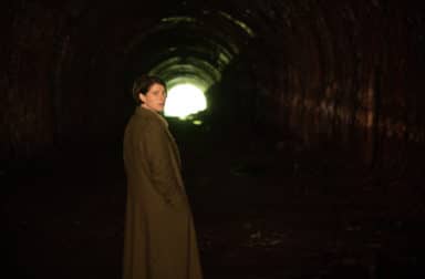 Jessie Buckley stands at the mouth of a tunnel, looking towards the camera. The end of the tunnel can be seen in the distance