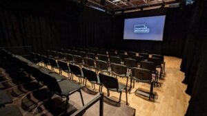 Interior view of the studio theatre with rows of black chairs facing a screen that displays the Taunton Brewhouse logo. The room has wooden flooring, black curtains, and a simple lighting rig overhead.