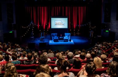 An interior view of the Auditorium taken from the back of the theatre. It is a full house. The stage is set up with a screen and two seats next to each other.